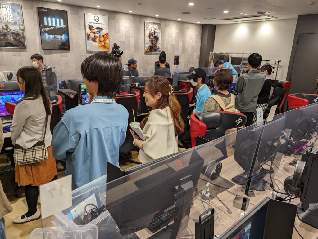 ゲーミングPCと椅子が並んでおり、画面に映るゲームの様子を10人ほどが立ち見している。