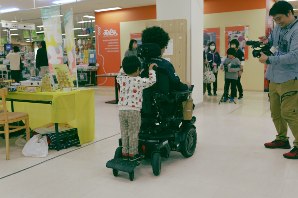 アフロの車椅子に連結された台に載る男の子