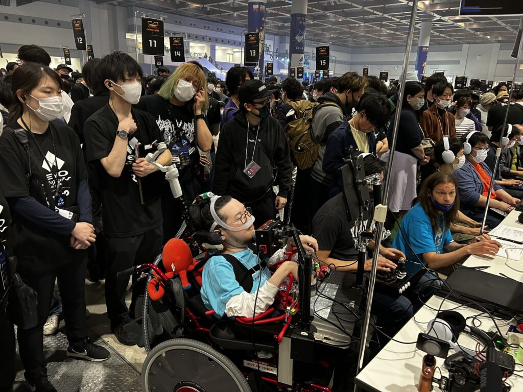 車椅子のJeniが他のプレイヤーと並んでゲームをプレイする様子。後ろにはスタッフや観客など大勢の人が立っている