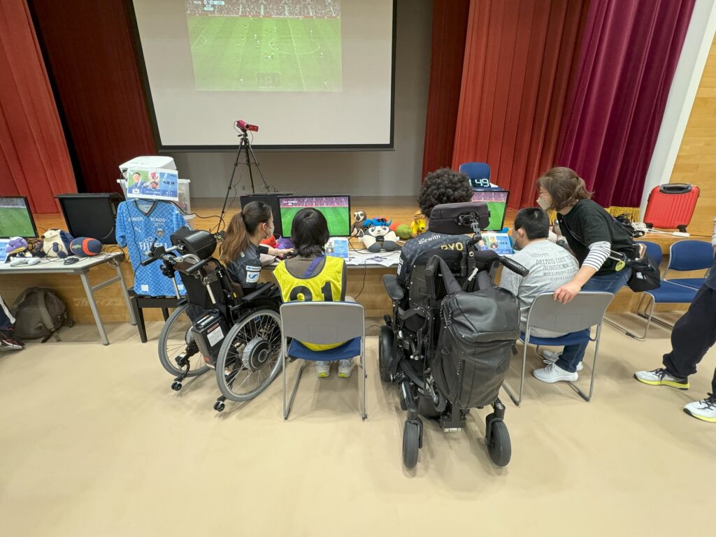 車椅子に乗ったイーパラユナイテッドのメンバーが隣に座った参加者と一緒にサッカーゲームをする様子
