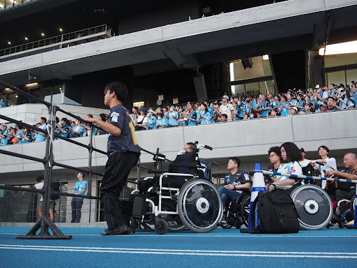 車いすメンバーがスタジアムでスポーツ観戦する様子。1人は車いすから立ち上がり、フェンスを持って立っている。