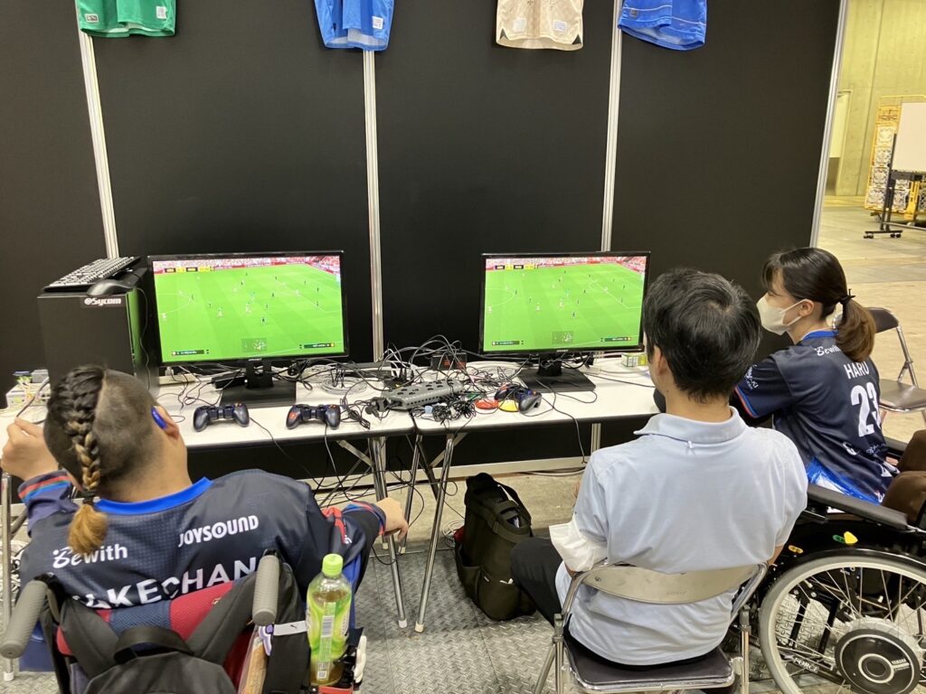 サッカーゲームが表示されたディスプレイを前にして座るイーパラユナイテッドのメンバー2人と一般来場者1人