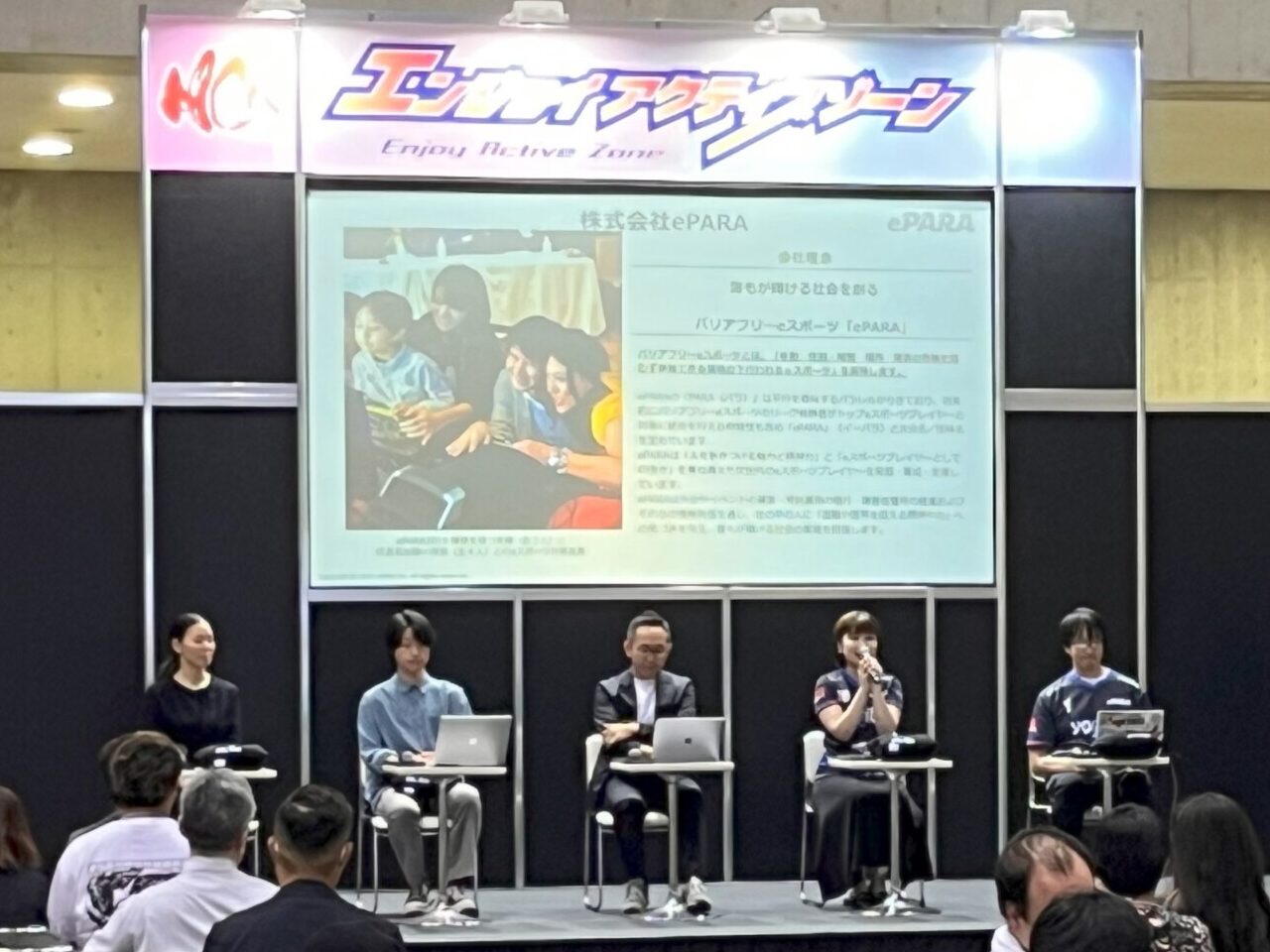 ステージ上に5人が並んで座り、うち1名がマイクを持って話している。登壇者の背後の画面には、株式会社イーパラを紹介する画像が写っている。