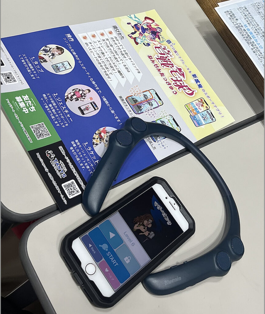 音だけで卓球のようなスポーツを楽しめるアプリゲーム「音戦宅球」のスマホ画面と、パンフレット、首にかける専用の音響デバイスの写真