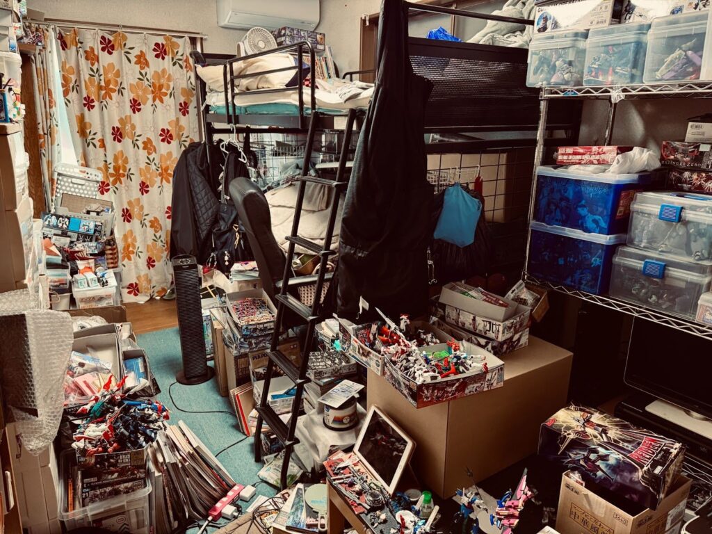 ロフトベッドの下や部屋中の収納ボックスにたくさんのプラモデルが収納されている。ごくわずかなスペースの残る作業台にiPadが立てかけられている。