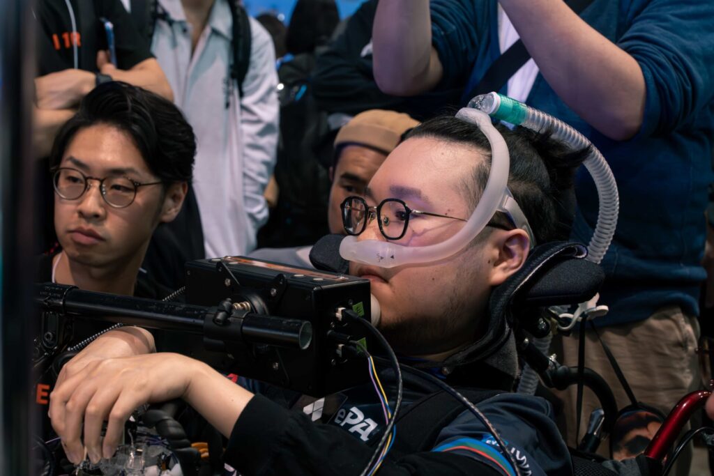 コントローラーを装着した車いすでゲームに臨むをプレイするJeniと、周りを取り囲む支援者や観戦者