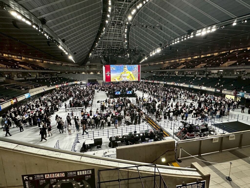 広大な体育館に多くの人がいる様子。会場中央に天井から吊るされた大画面にストリートファイター6のキャラクターが映っている
