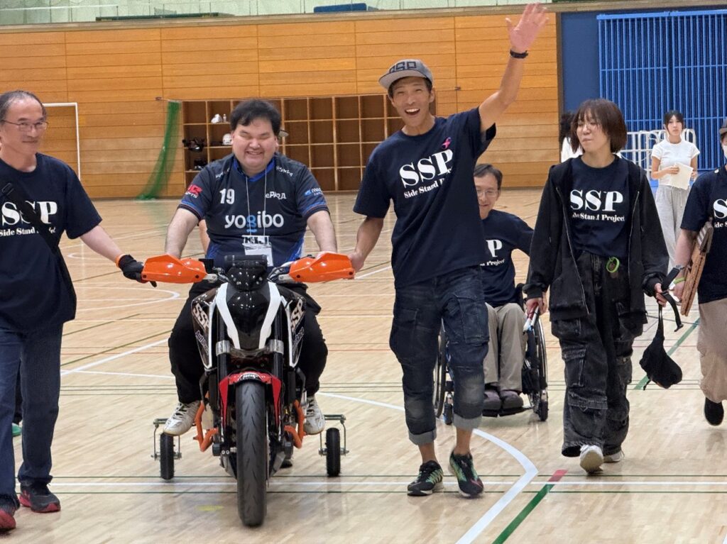 補助輪付きバイクにまたがる笑顔の直也と周りでサポートするスタッフ