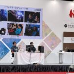 ステージ上に座り障害当事者のeスポーツプレイヤーのスライドを背に事業紹介する加藤代表とNAOYA選手の写真
