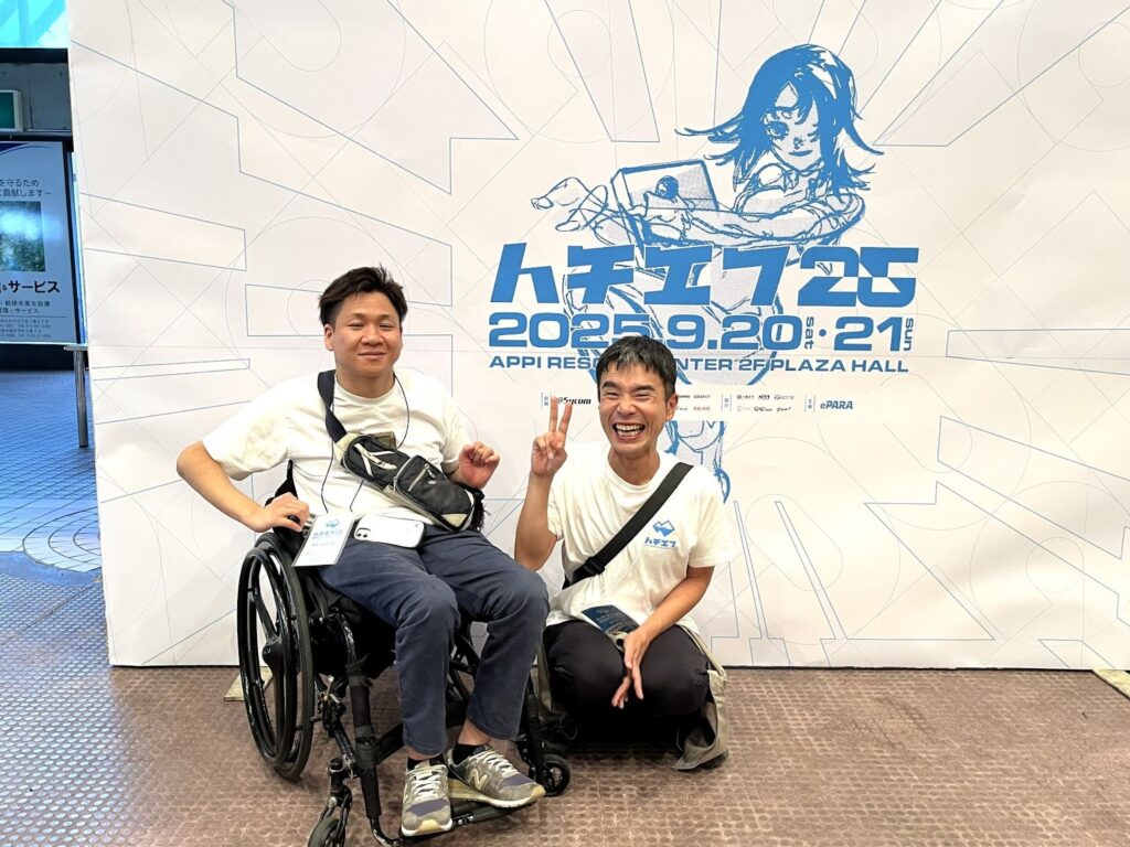 ハチエフ25のキービジュアルパネルの前で、微笑む車椅子のゆうたろうさんとピースサインで笑顔のさなけんさん