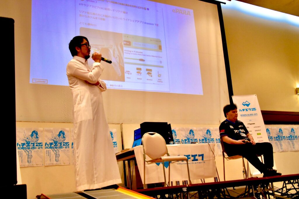 ステージでサウジアラビアの正装（白の丈の長いシャツワンピース型の衣装）を着た加藤代表が立ってプレゼンを行っている様子。並んでNAOYA選手が座っている。