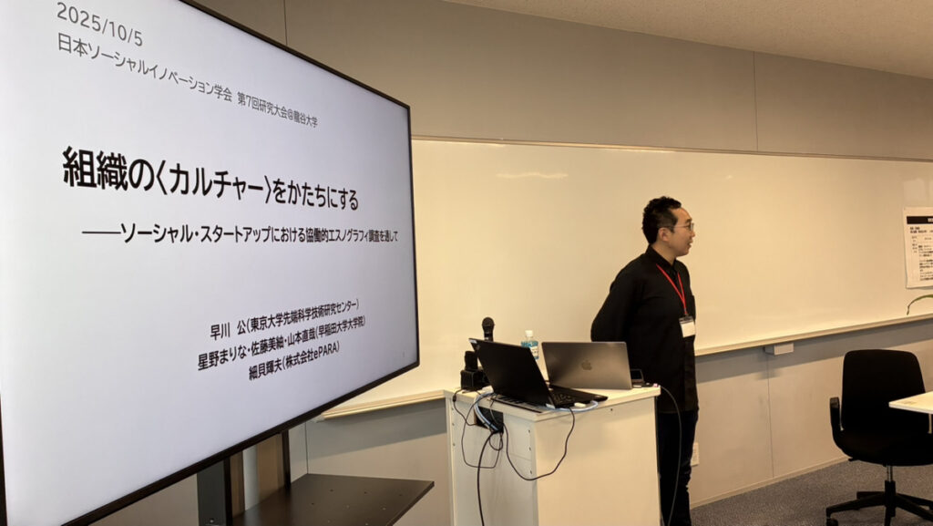教室の前に立って話す早川氏。横に置かれた大きい画面に「組織のカルチャーをかたちにする　ソーシャル・スタートアップにおける協働的エスのグラフィ調査を通して」という研究タイトルと研究参加者名が表示されている