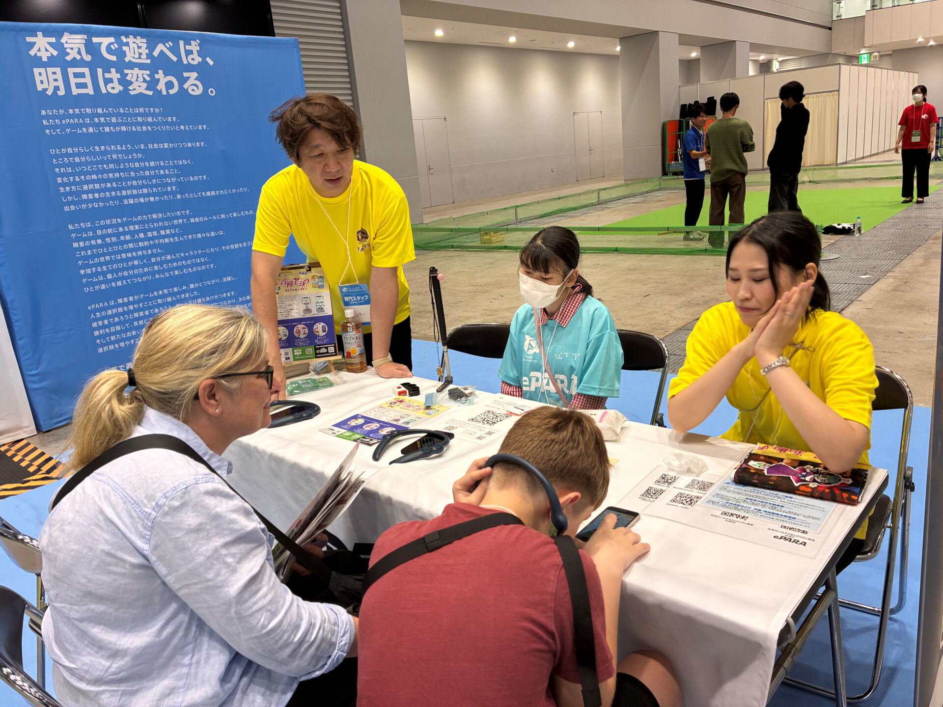 黄色のTシャツを着たタニーズの開発スタッフ二名と全盲プレイヤーのゆきさんが、長机を挟んで外国人の参加者に音戦宅球のやり方を教えている様子