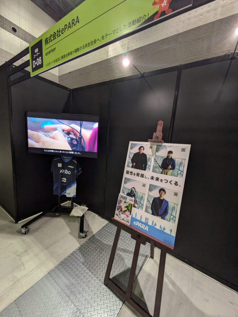 イーパラのブース。会社のポスターが飾られ、ブース内に置かれたディスプレイにゲームコントローラーを操作する様子が映し出されている。ユニフォームも飾られている。
