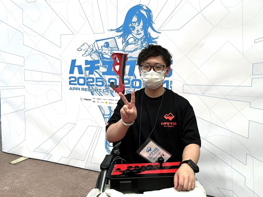 ハチエフ25のキービジュアルパネルの前でピースサインをする車椅子のまるぽすさん。膝の上にはコントローラー。
