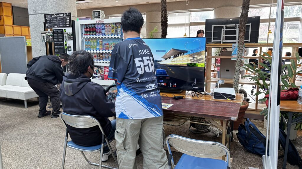 レーシングゲームブースで体験者に説明をするジューク