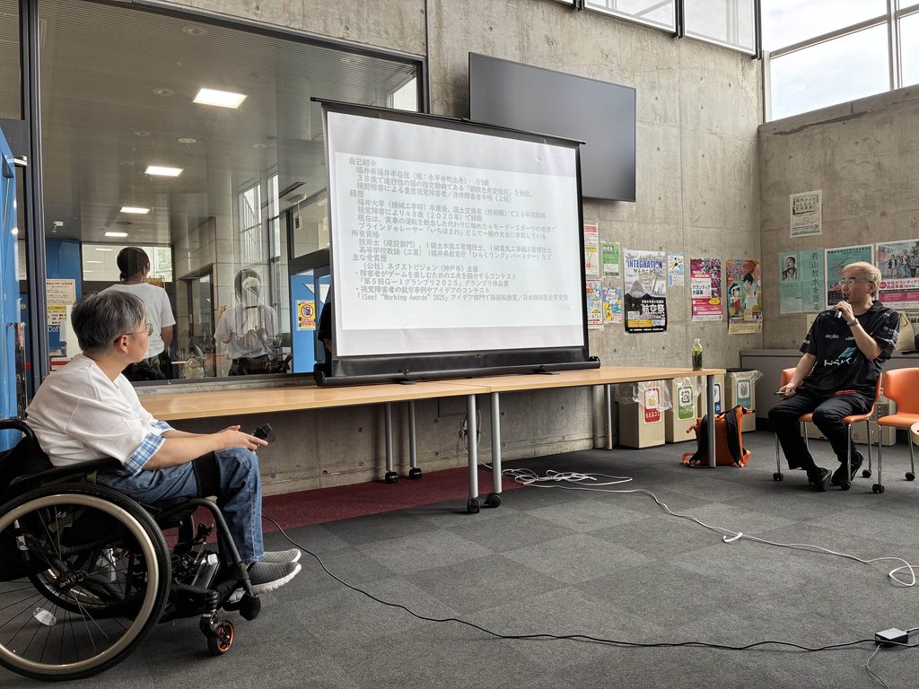 室内で行われている講演の様子。手前にスクリーンのスライドを見る車いすの参加者、右側にマイクを持って話す登壇者の男性が写っている。